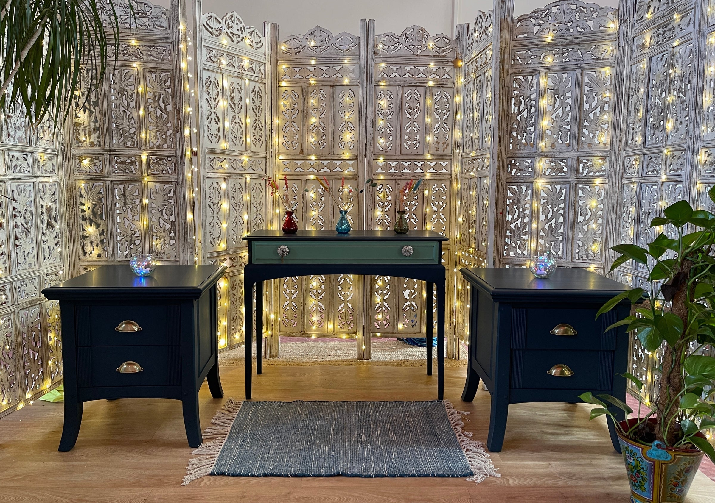 Two small nay painted cabinets with a console table and a carved indian wood background with fairy lights, a small rug and some plants