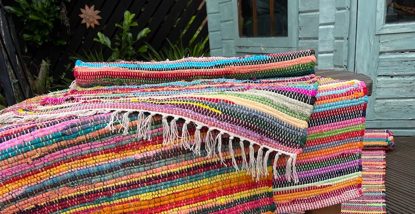 A pile of rag rugs lying on a small table sitting on garden decking with a pale turquoise garden room in the background