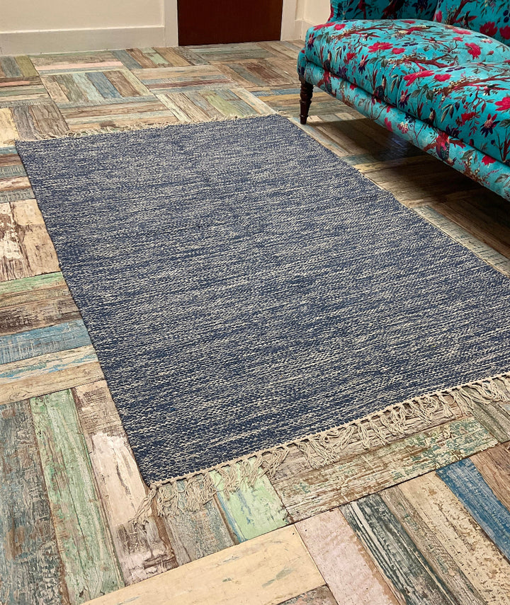Blue handwoven cotton rug with fringe edges, styled in eclectic room with patterned floor and floral sofa.