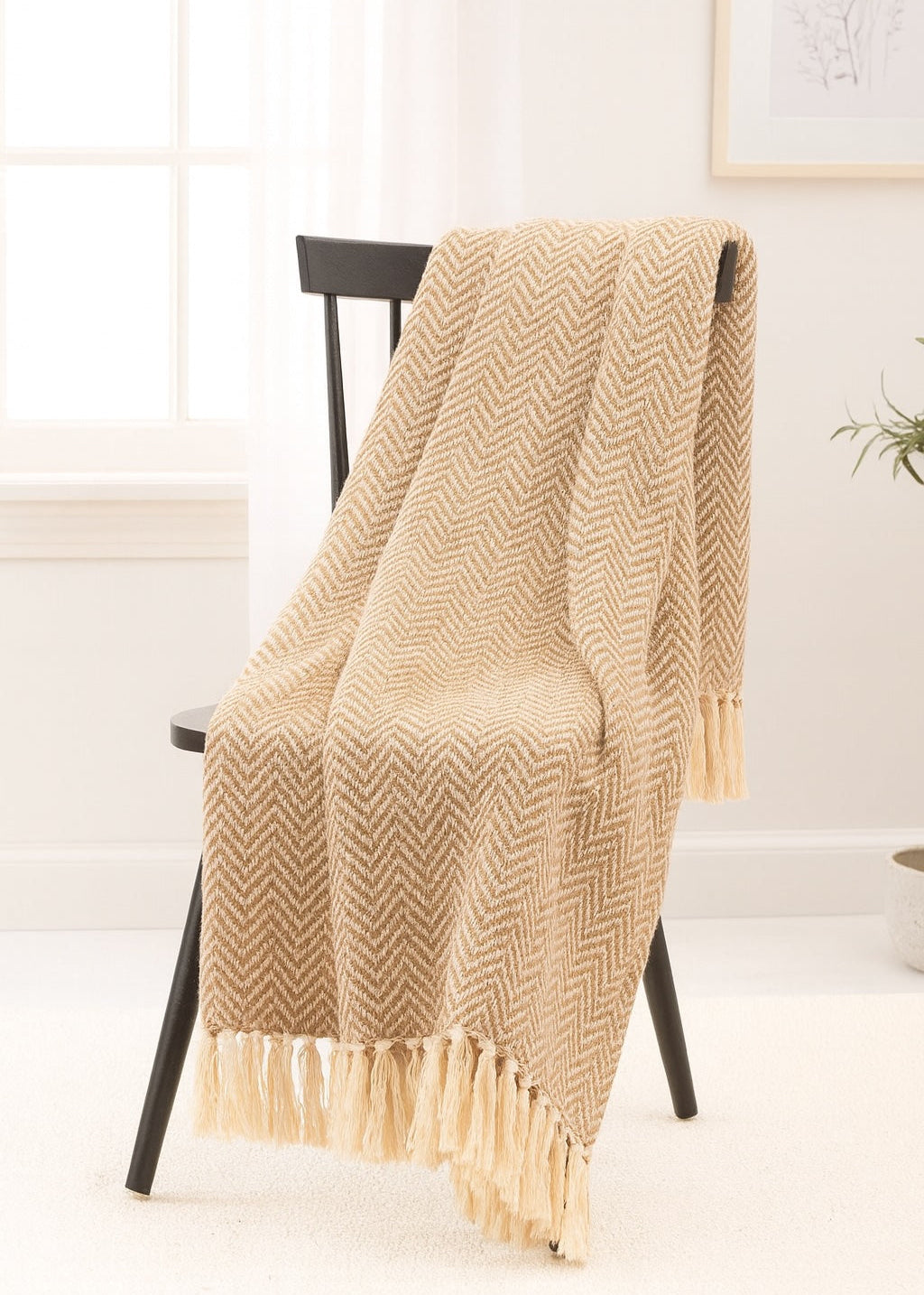 Beige herringbone patterned throw blanket draped over a chair in a room with a white wall and plant.