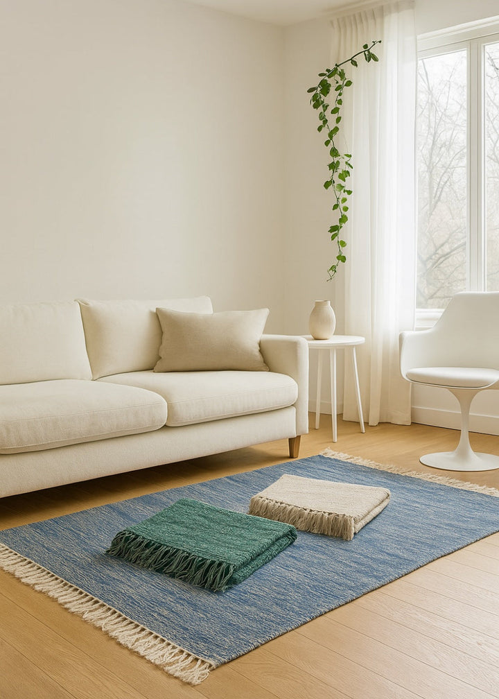 Blue cotton rug with grey and green folded rugs in a minimalist living room with cream sofa and wood flooring.