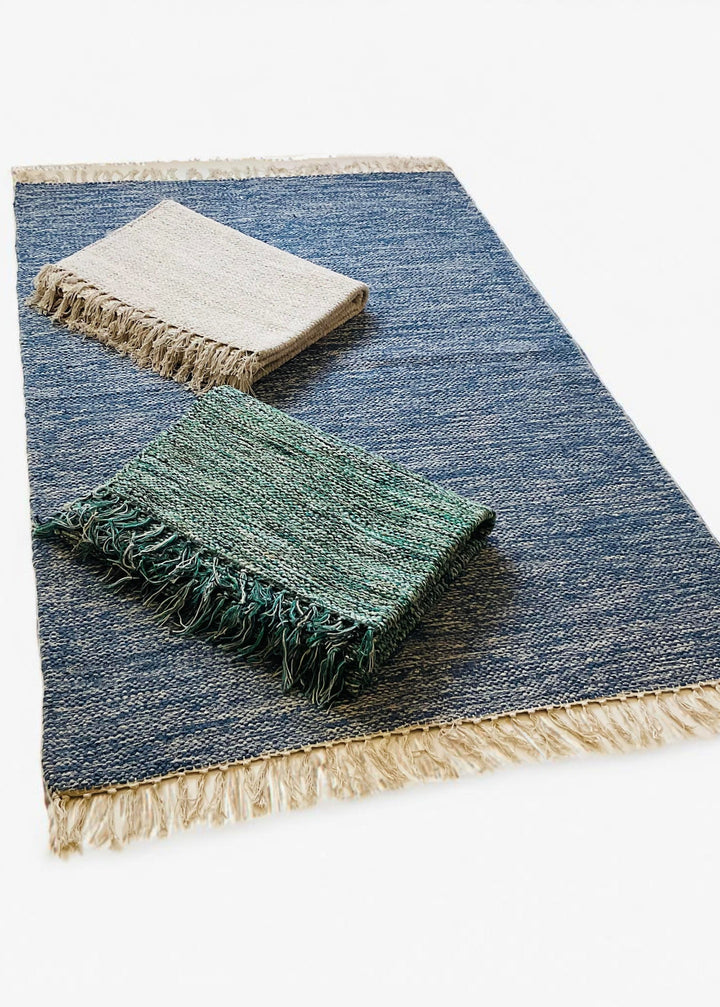 Drift Rug made from cotton yarn, shown in blue with two folded colour options—grey and green—All rugs feature a natural fringe detail.