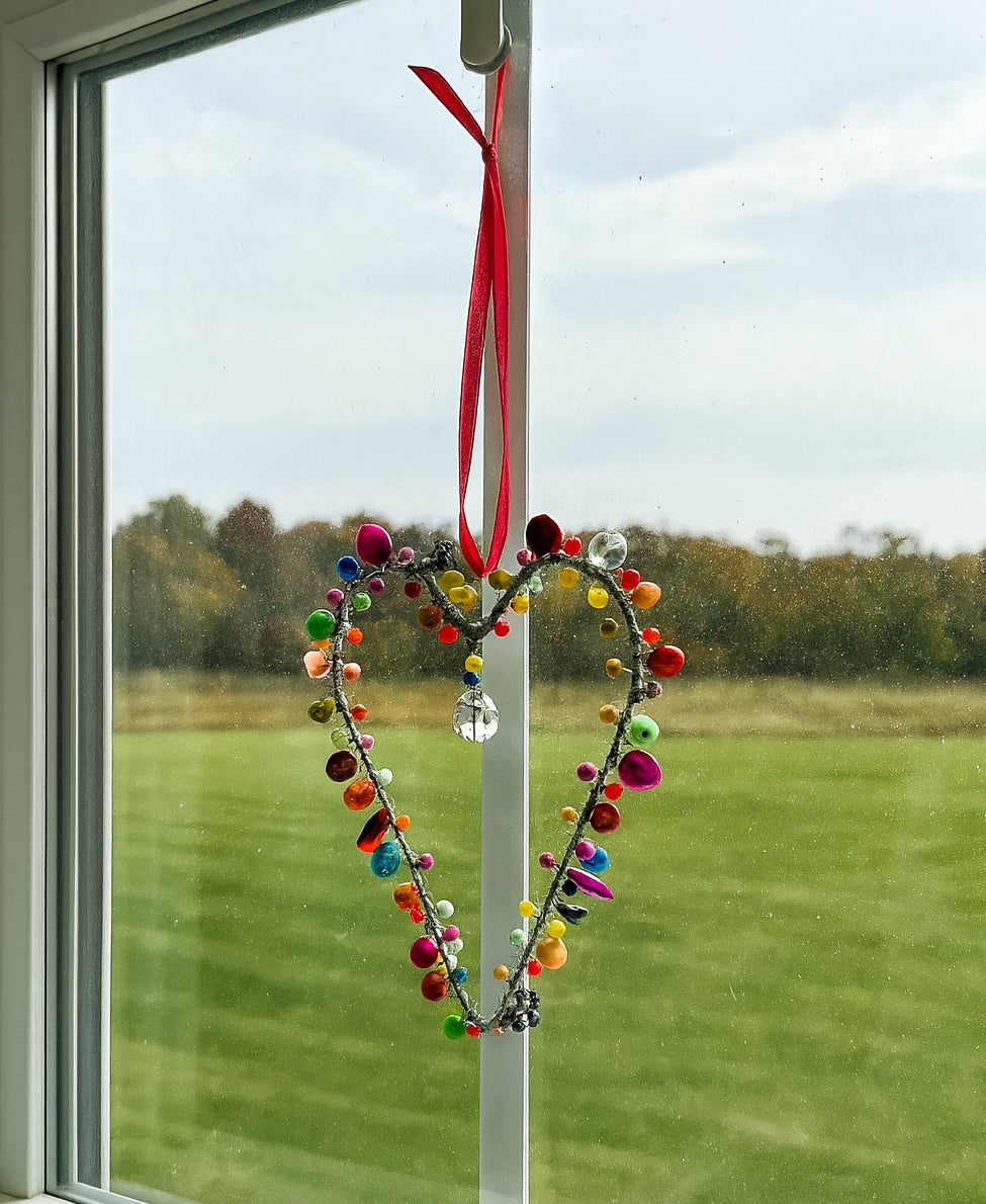 Colorful heart-shaped decoration hanging on a window with a view of a grassy field and trees.
