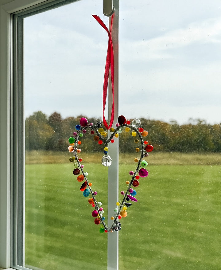 Colorful heart-shaped decoration hanging on a window with a view of a grassy field and trees.