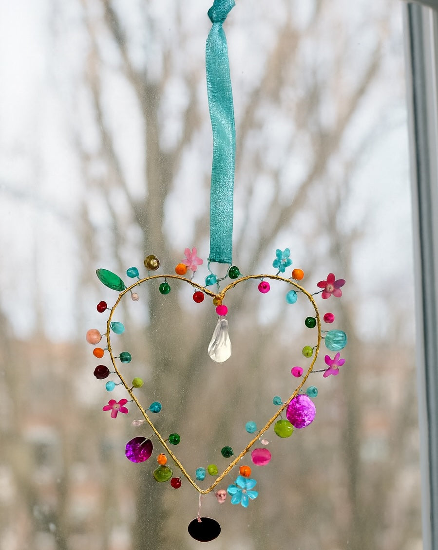 Decorative heart-shaped hanging ornament with colourful beads and flowers against a blurred natural background.