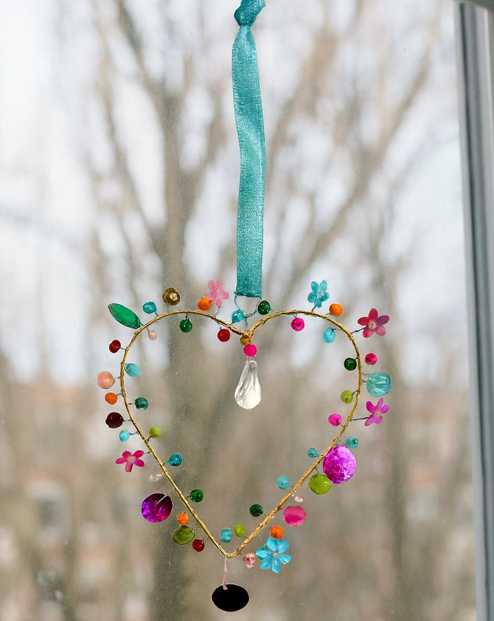 Decorative heart-shaped hanging ornament with colourful beads and flowers against a blurred natural background.