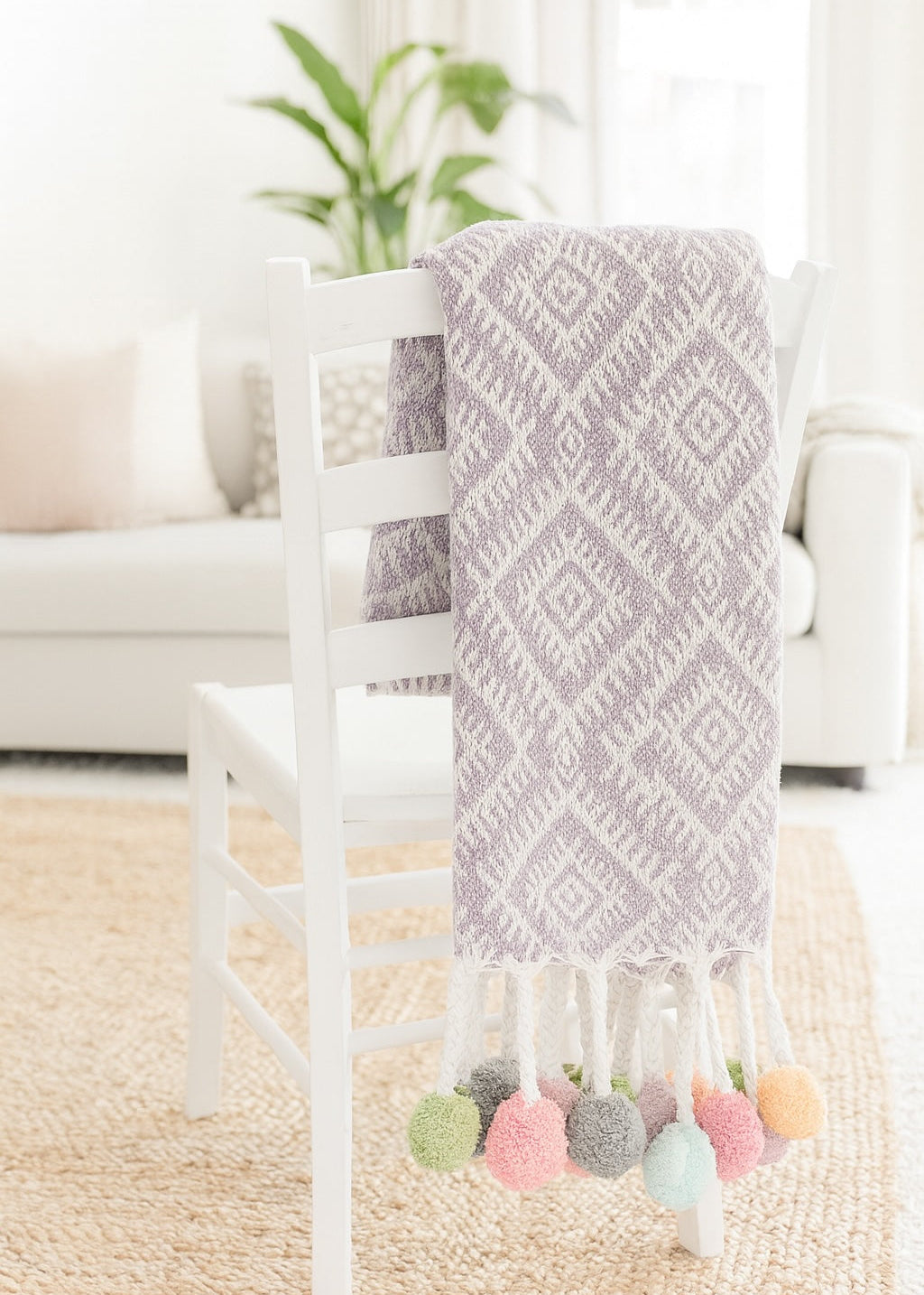 Lilac patterned blanket with colourful pom-poms draped over a white chair in a bright room.