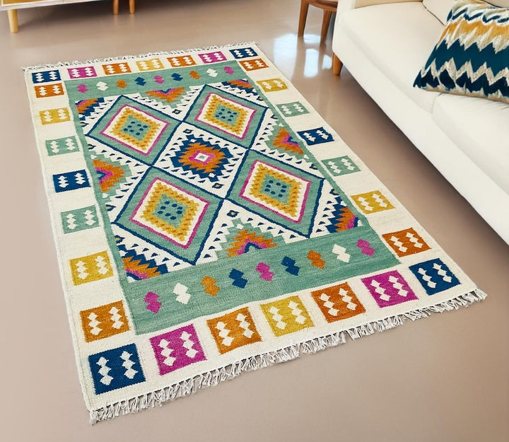 Colourful Indian kilim rug with green and blue geometric pattern and fringe, displayed in a modern room beside a cream sofa and patterned cushion.