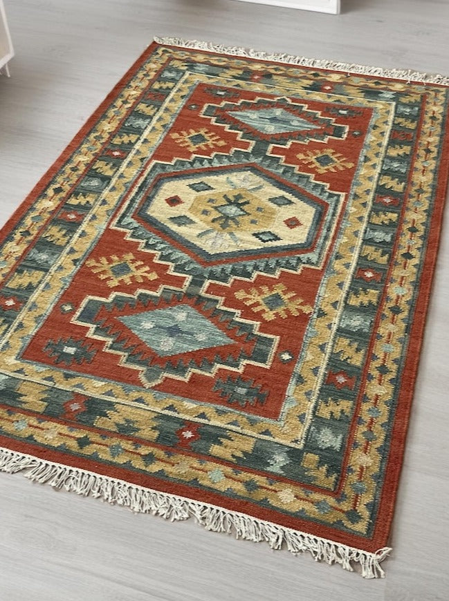 Flatweave wool-cotton kilim rug with geometric pattern in rust, gold, green, and cream on light wood floor.