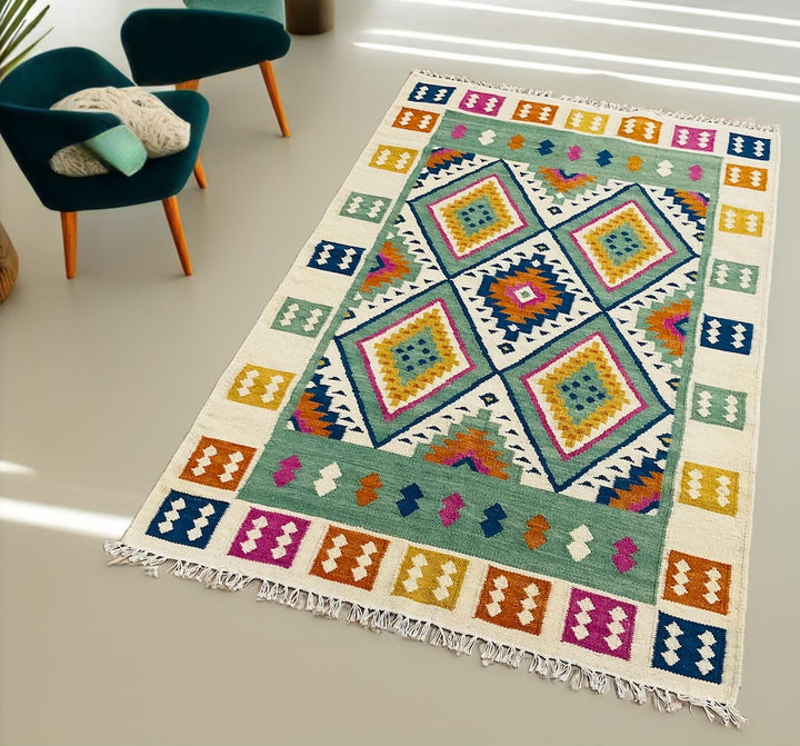 Multicolour handwoven Indian kilim rug with geometric diamond pattern and fringe, shown in a modern room with teal chairs and neutral flooring.