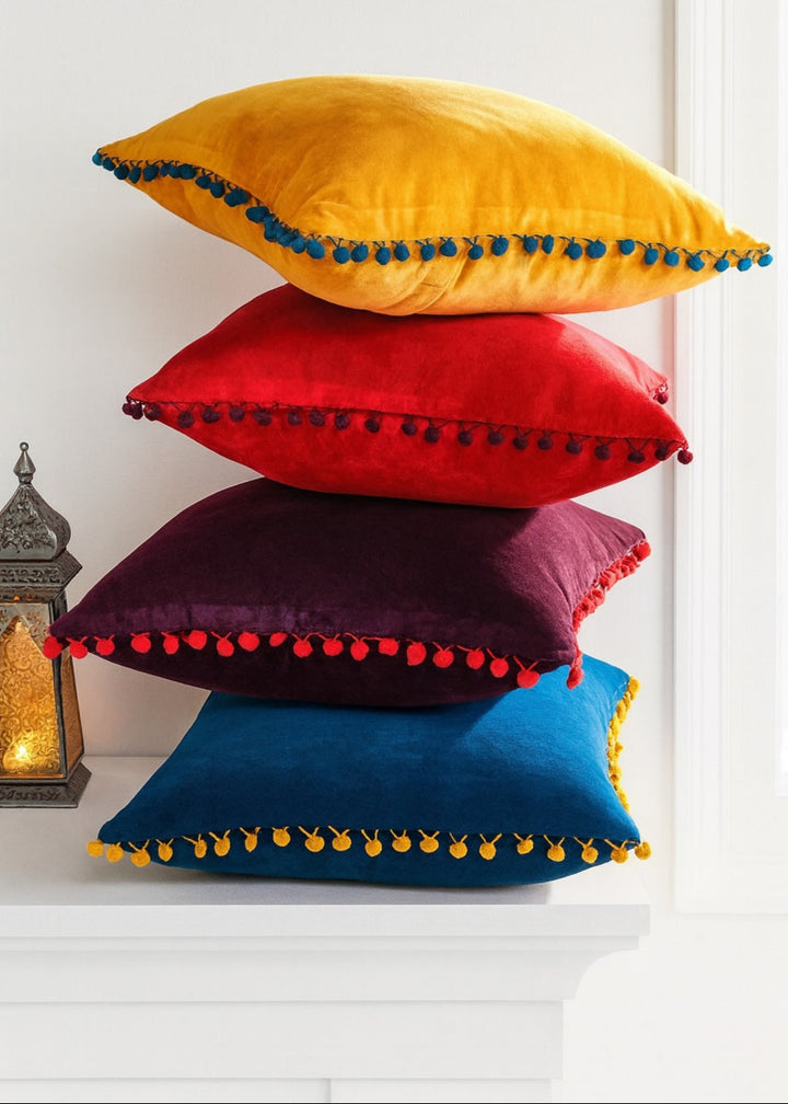Stack of colourful pillows with pom-pom trim on a white surface.