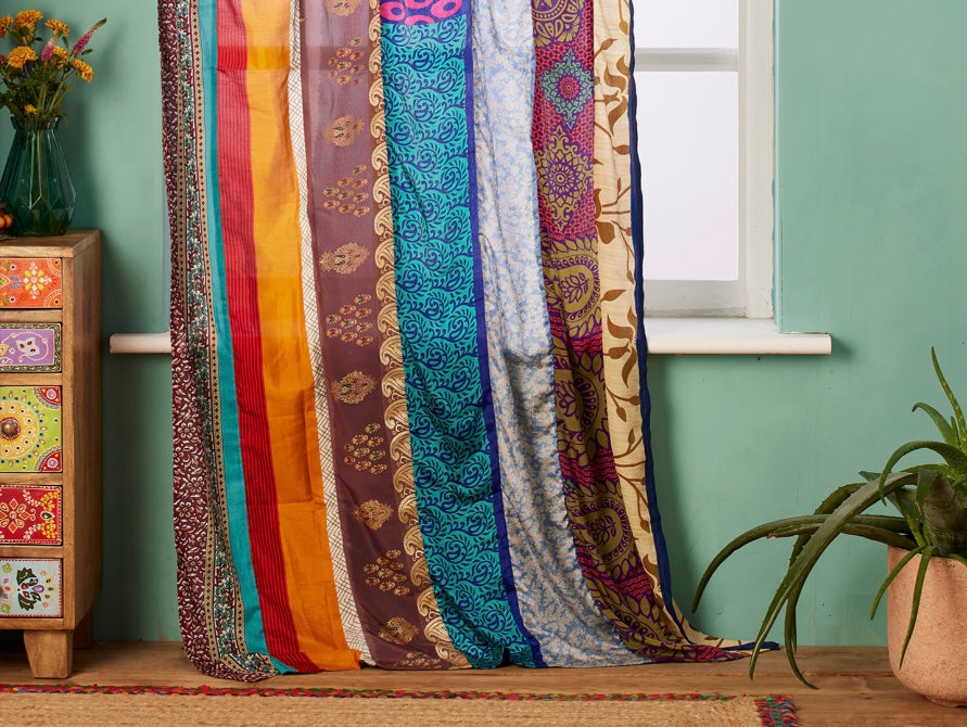 A pair of voile curtains made with multi coloured sari fabric hanging from a window with a bohemian chest of drawers and a large green plant on the floor.