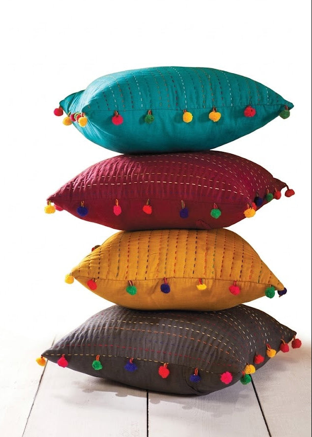 Four decorative throw pillows stacked vertically on a white wooden floor against a white background. The pillows are teal, burgundy, mustard yellow, and dark gray, each decorated with colorful tassels along the edges.