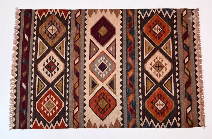 A square woven kilim rug with a geometric tribal pattern in shades of brown, rust red, beige, and black, featuring fringe on the sides.