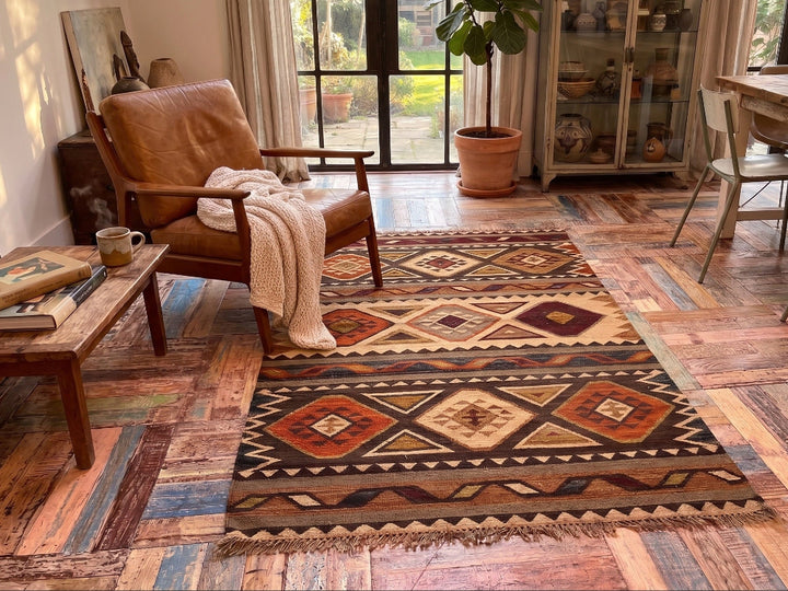 A rectangular Southwestern-style area rug with geometric diamond patterns in brown, beige, orange, and red, styled on a rustic wood floor in a cozy living room setting.