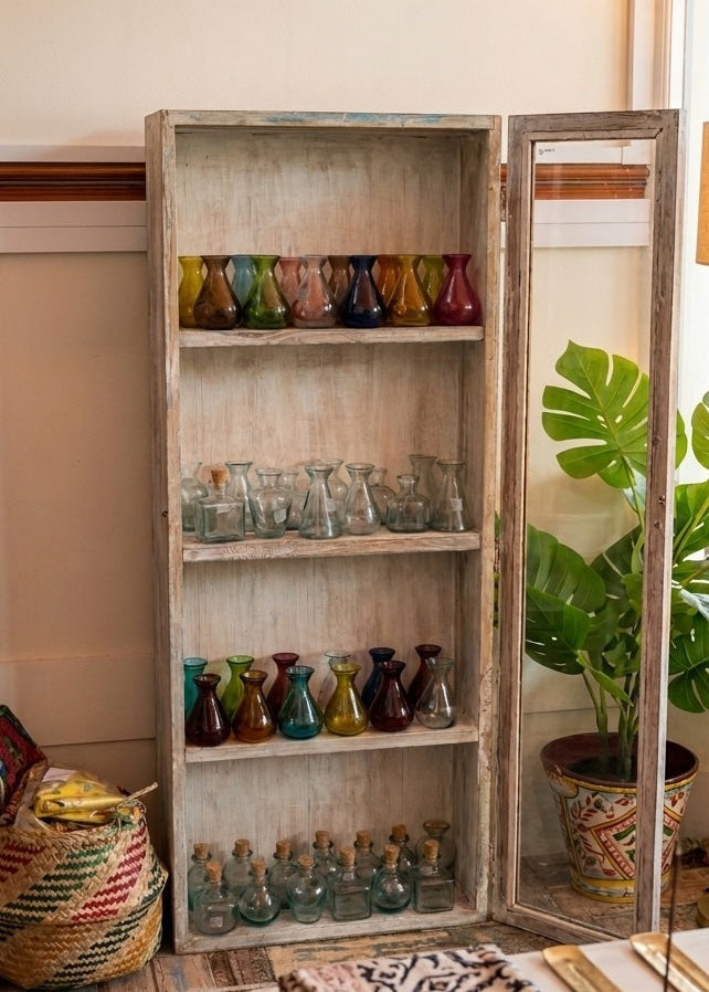 A rustic, weathered wooden display cabinet with a mirrored door stands in a styled room, filled with colorful glass vases and small bottles. A potted green plant sits to the right of the open cabinet, and patterned textiles are draped on a table in the foreground.