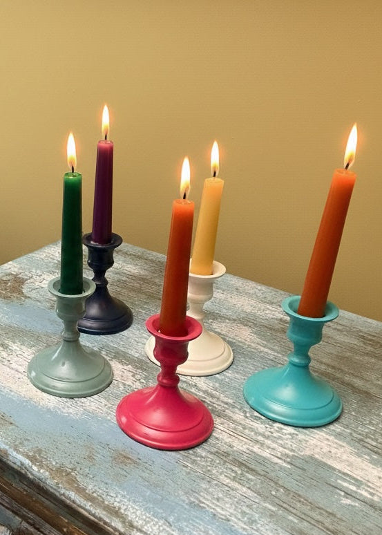 Colourful candles in various candlesticks on a wooden surface with a beige wall background.