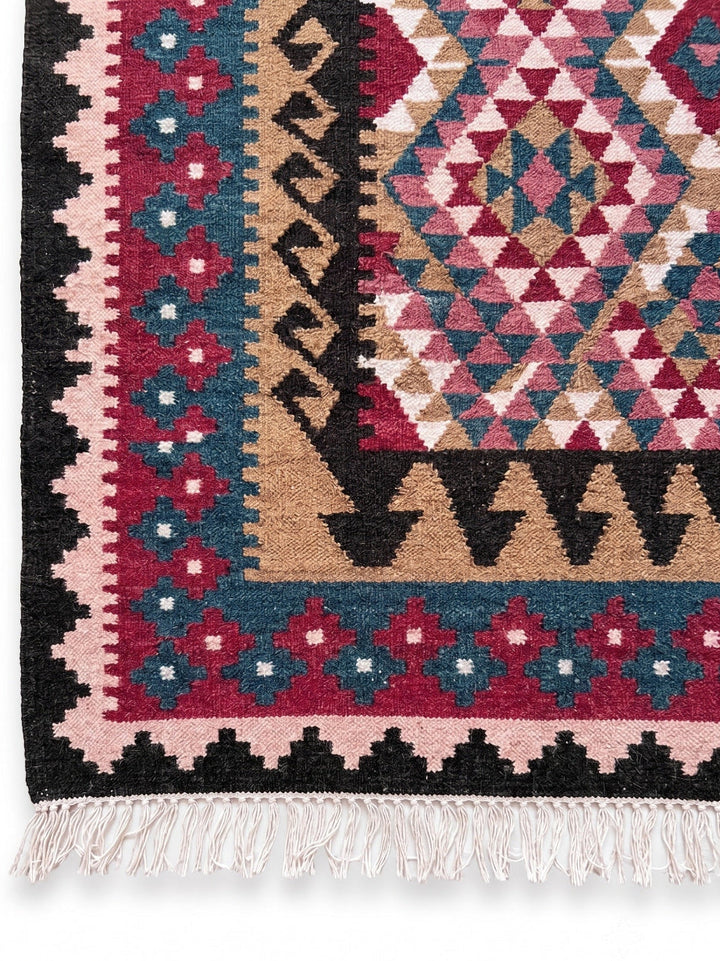A close-up of a patterned area rug showing its corner, featuring geometric tribal designs in pink, beige, teal, black with white fringe.