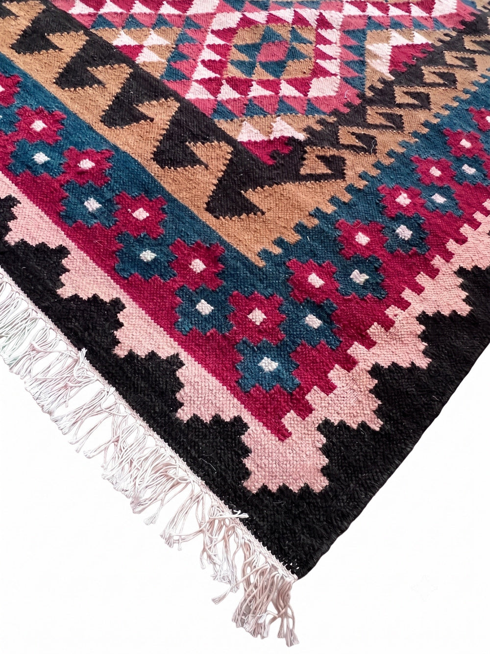 A close-up of a woven rug corner showing a geometric pattern with black, pink, teal blue, and tan bands, finished with white fringe on a white background.