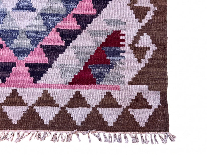 Close-up of a woven geometric rug corner showing pink, brown, cream, blue, and burgundy triangular patterns with fringe along the bottom edge, photographed on a white background.