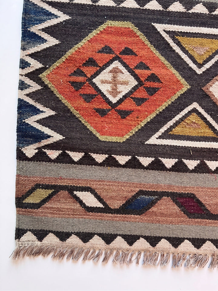 A close-up of a woven geometric rug showing a central orange-red diamond motif with black, cream, and olive accents, surrounded by navy, beige, and brown bands with fringe at the bottom.