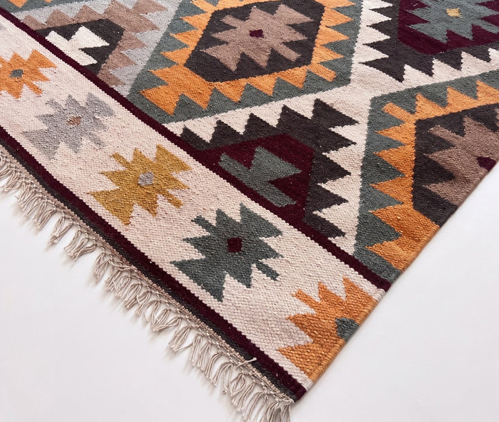 A close-up of a woven geometric area rug with fringe, featuring a Southwestern-style pattern in cream, orange, dark red, gray, and black on a white background.
