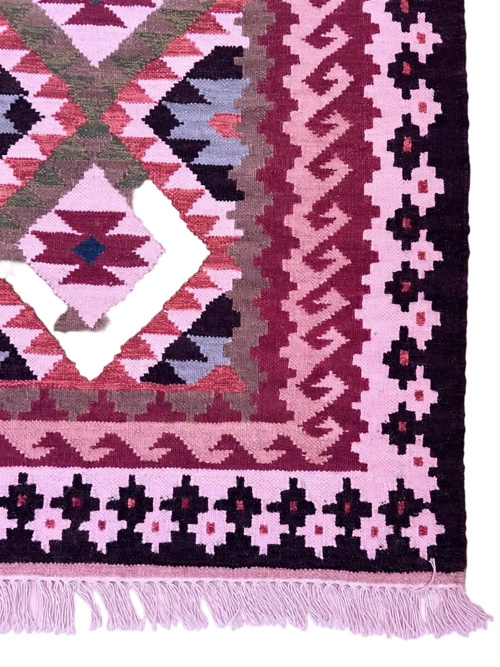 A close-up of a patterned rug corner showing geometric diamond motifs and a decorative border in pink, burgundy, black, olive green, and light grey, with matching pink fringe along the bottom edge.