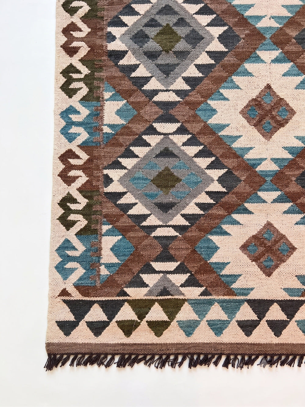 A close-up of a woven geometric rug corner showing a cream base with brown, teal blue, gray, and olive green diamond and triangle motifs, finished with dark fringe.