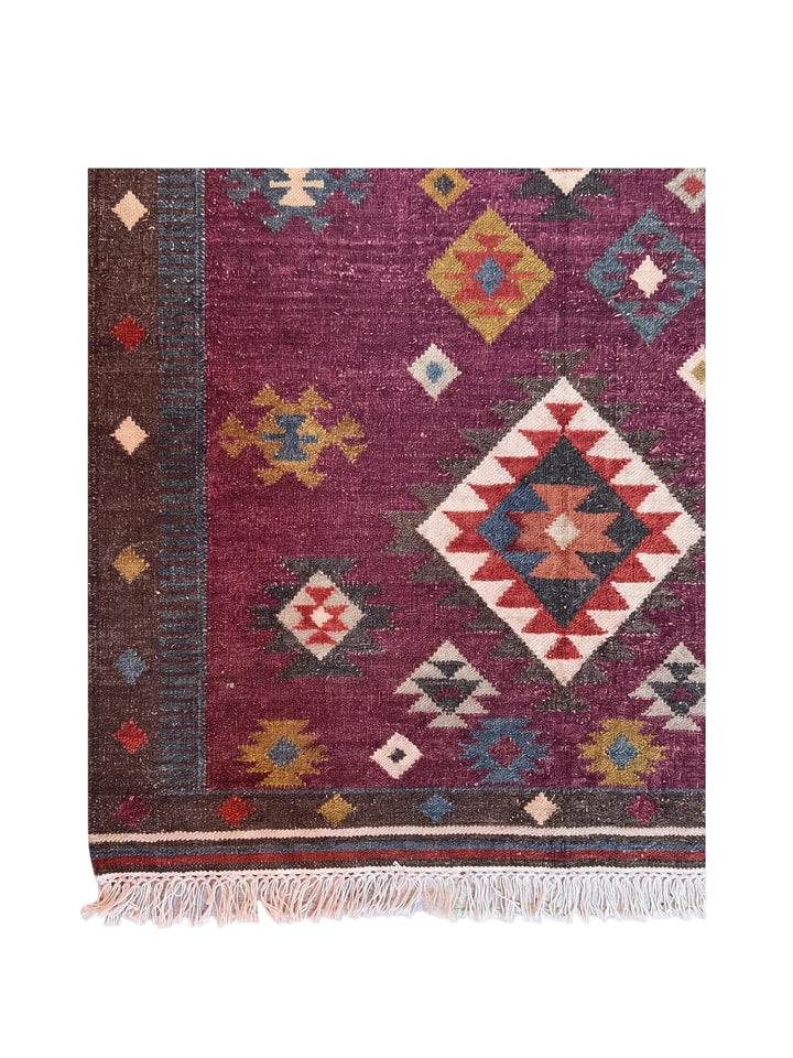 A square kilim-style area rug with a deep purple field, featuring a large central diamond motif and smaller geometric accents in red, blue, beige, and yellow. The rug has a dark brown border with small diamond motifs and a fringed edge.