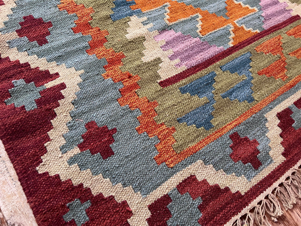 A close-up of a woven kilim rug with a geometric pattern in muted shades of blue, rust orange, burgundy, cream, and olive green, featuring a fringed edge.