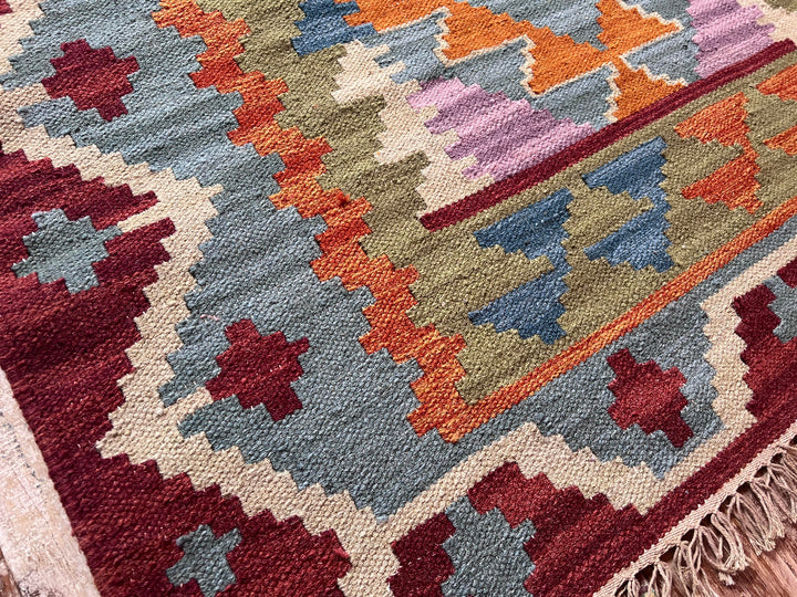 A close-up of a woven kilim rug with a geometric pattern in muted shades of blue, rust orange, burgundy, cream, and olive green, featuring a fringed edge.