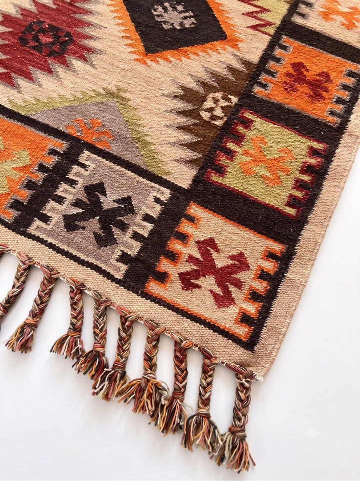 A close-up of a woven kilim rug with a Southwestern geometric pattern in beige, brown, orange, red, and green, featuring a fringed edge.