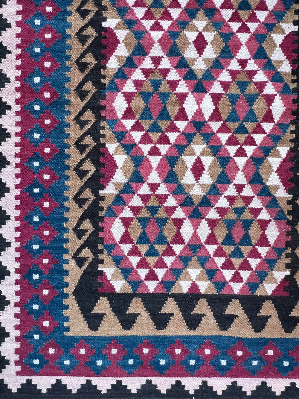 A close-up of a woven kilim rug featuring a geometric diamond pattern in pink, blue, beige, and cream, with a decorative border.