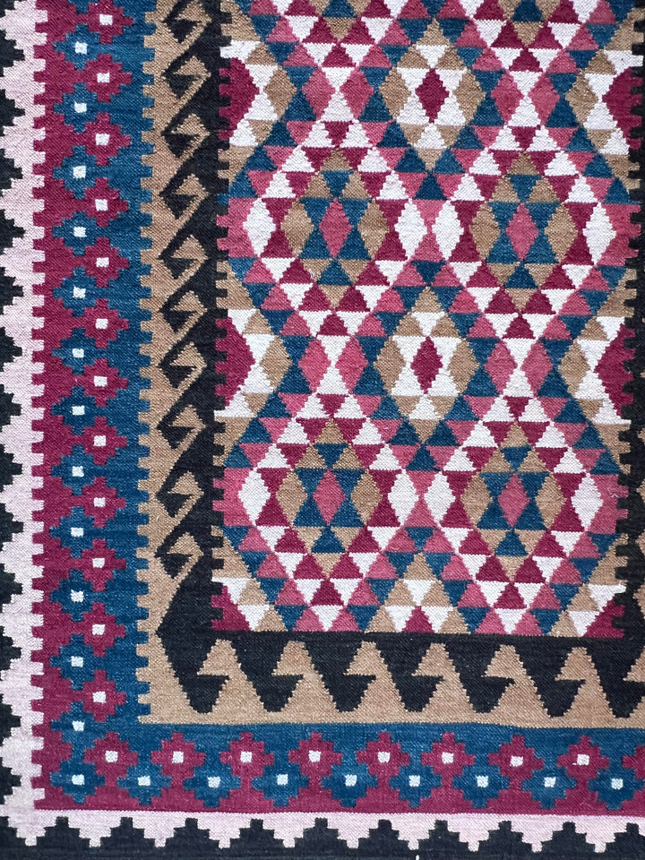A close-up of a woven kilim rug featuring a geometric diamond pattern in pink, blue, beige, and cream, with a decorative border.