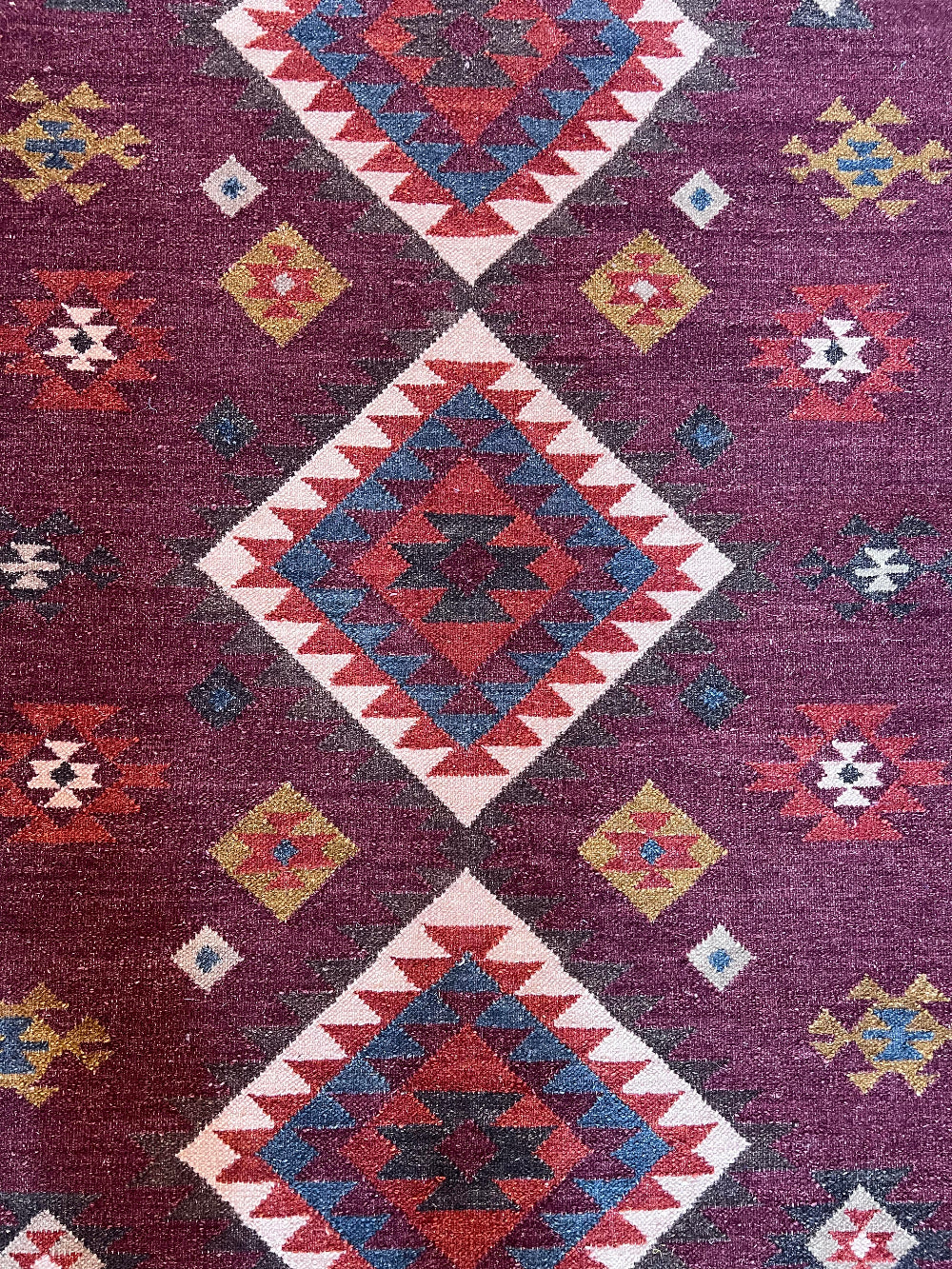 A close-up of a woven rug with a deep purple field and large diamond motifs in red, cream, blue, and yellow.