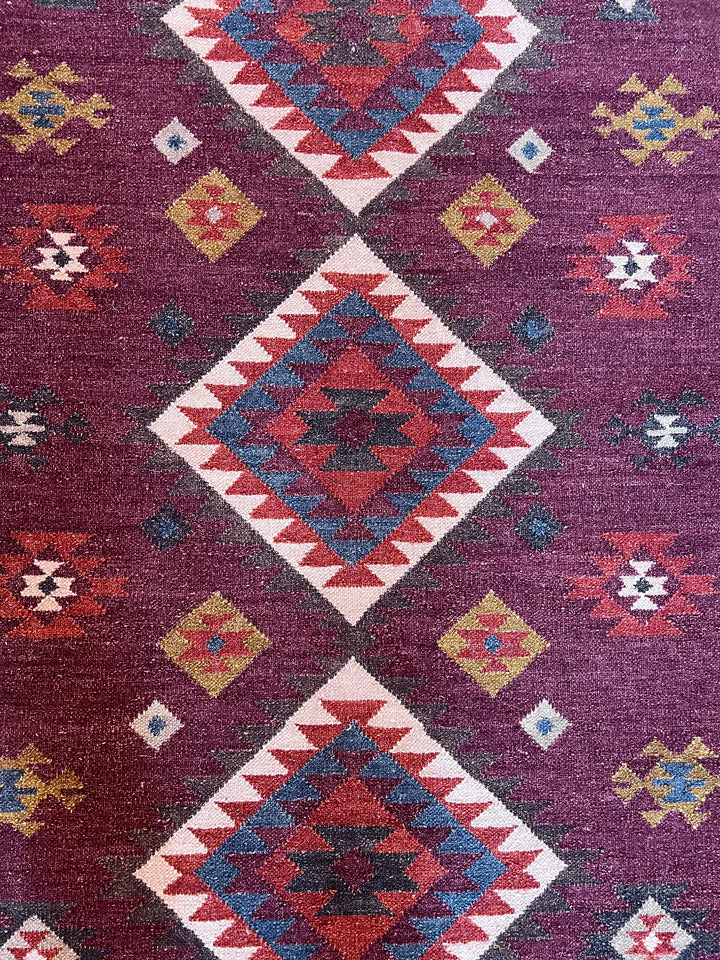 A close-up of a woven rug with a deep purple field and large diamond motifs in red, cream, blue, and yellow.