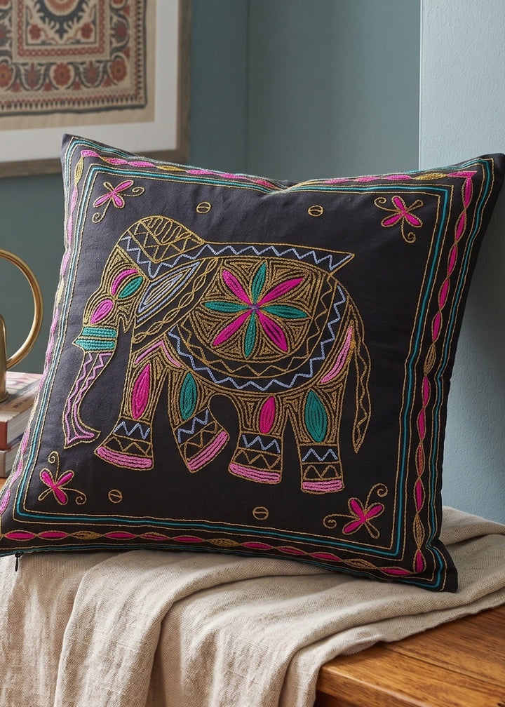 A square decorative throw pillow with a black background and a colourful embroidered elephant design, styled on a wooden bench in a home interior setting.