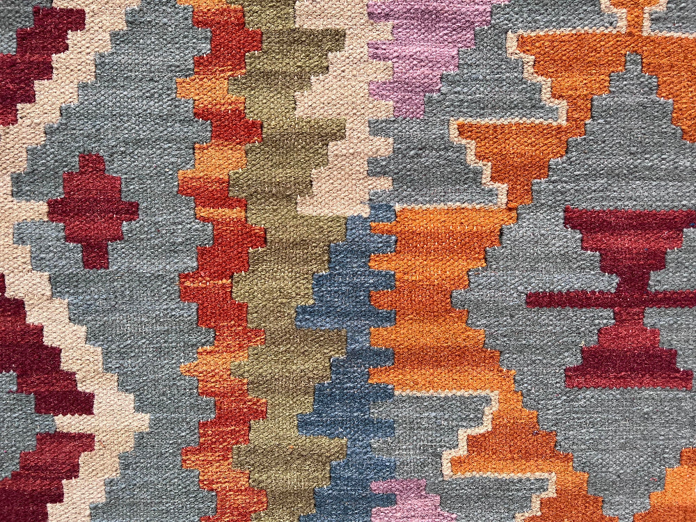 A close-up of a woven textile with a geometric tribal pattern in muted blue-gray, rust orange, cream, burgundy, olive, and lavender.