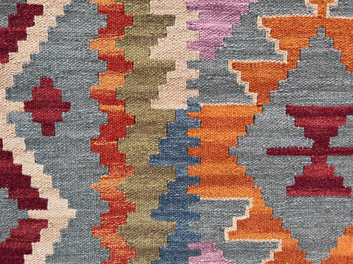 A close-up of a woven textile with a geometric tribal pattern in muted blue-gray, rust orange, cream, burgundy, olive, and lavender.
