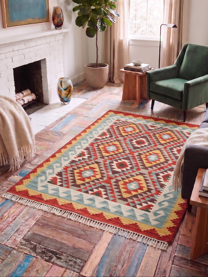 A colorful geometric area rug with fringe, styled in a cosy living room with a green armchair, a fireplace, and rustic wood floors.