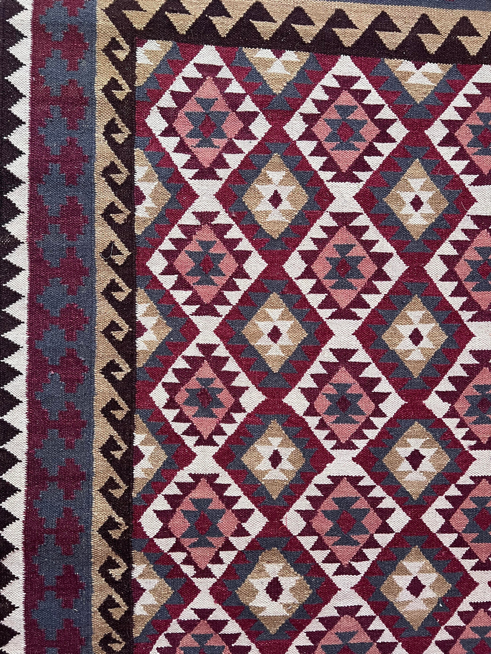 A close-up of a woven geometric rug with repeating diamond and triangle motifs in burgundy, cream, beige, and slate blue, plus a dark border.