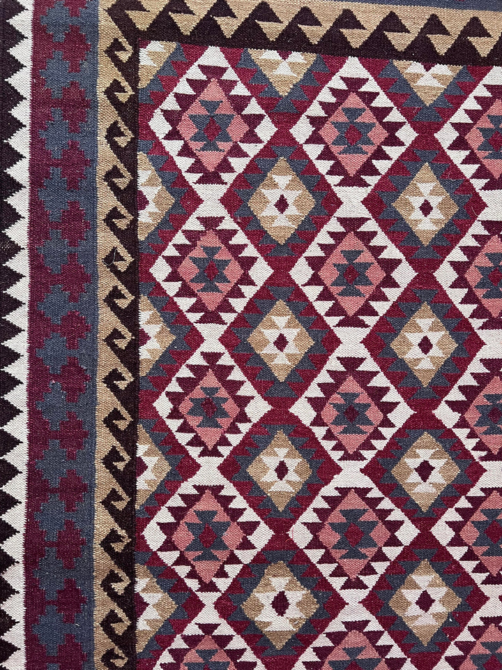 A close-up of a woven geometric rug with repeating diamond and triangle motifs in burgundy, cream, beige, and slate blue, plus a dark border.