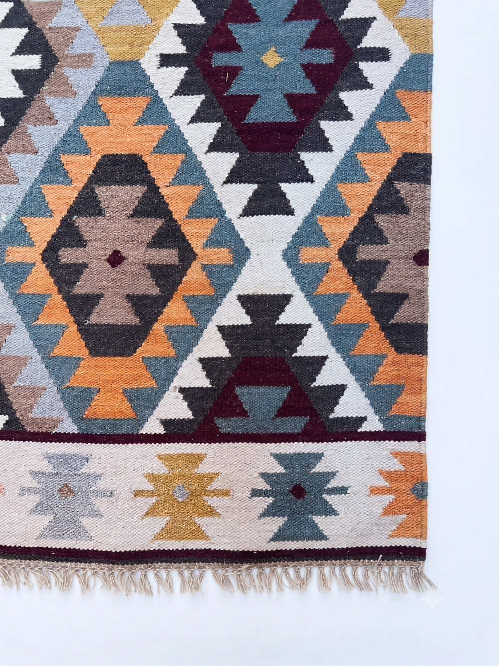A close-up of a woven geometric rug with a Southwestern-style pattern in orange, blue, beige, gray, and burgundy, featuring a fringed edge on a white background.