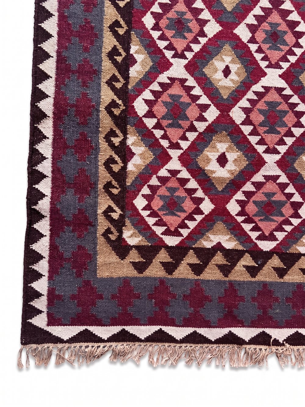 A close-up of a woven rug corner showing a geometric diamond pattern in burgundy, beige, pink, and dark gray, with a decorative border and tasseled fringe.