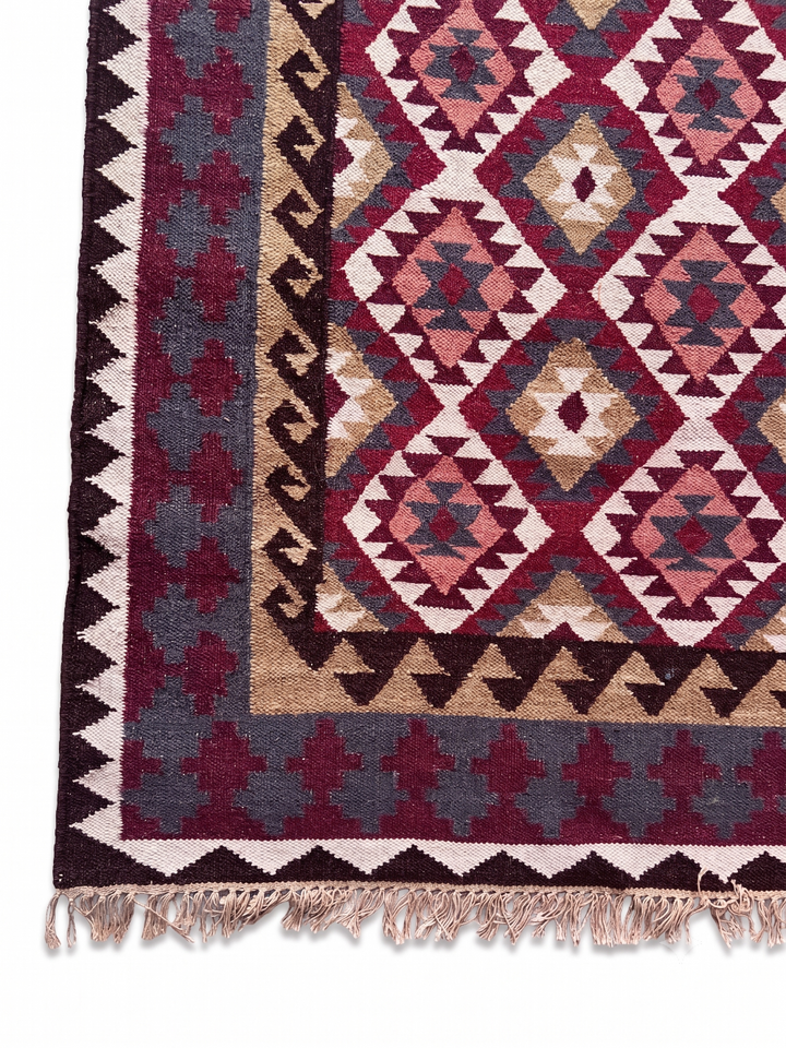 A close-up of a woven rug corner showing a geometric diamond pattern in burgundy, beige, pink, and dark gray, with a decorative border and tasseled fringe.