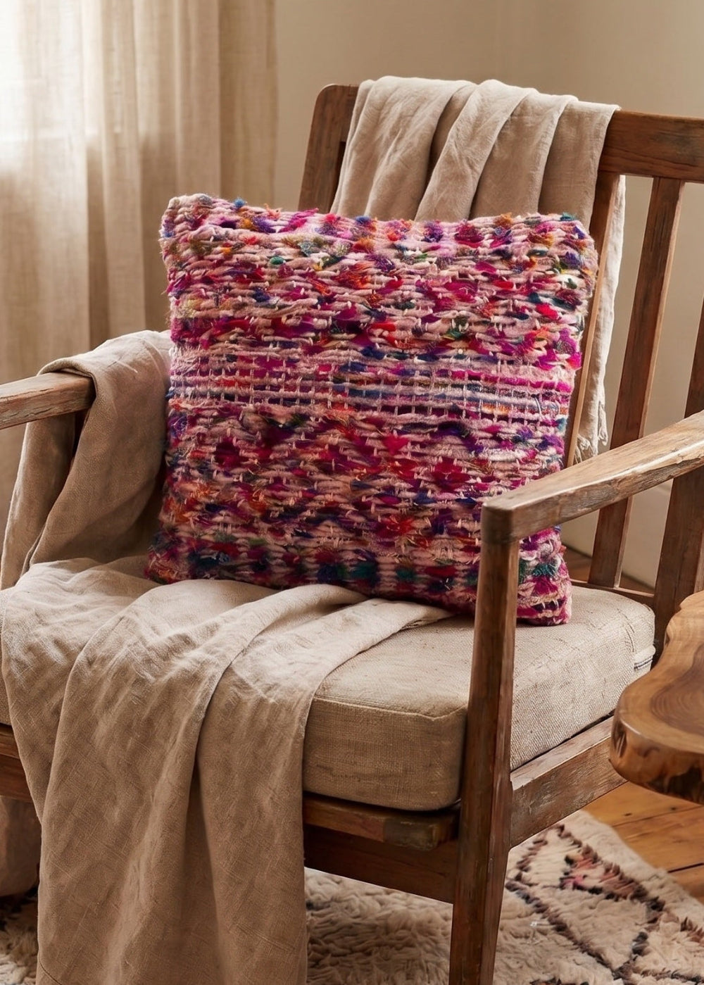 A rustic wooden armchair in a cosy room setting, styled with a beige linen throw blanket and a vibrant, multi coloured geometric patterned throw pillow. A matching patterned table lamp sits on a rustic wood side table next to the chair.