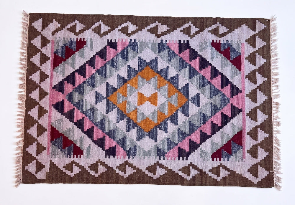 A woven kilim rug with a geometric diamond pattern in shades of brown, pink, navy, grey, and orange, featuring white fringe on the sides.