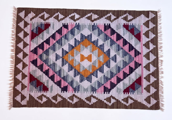 A woven kilim rug with a geometric diamond pattern in shades of brown, pink, navy, grey, and orange, featuring white fringe on the sides.