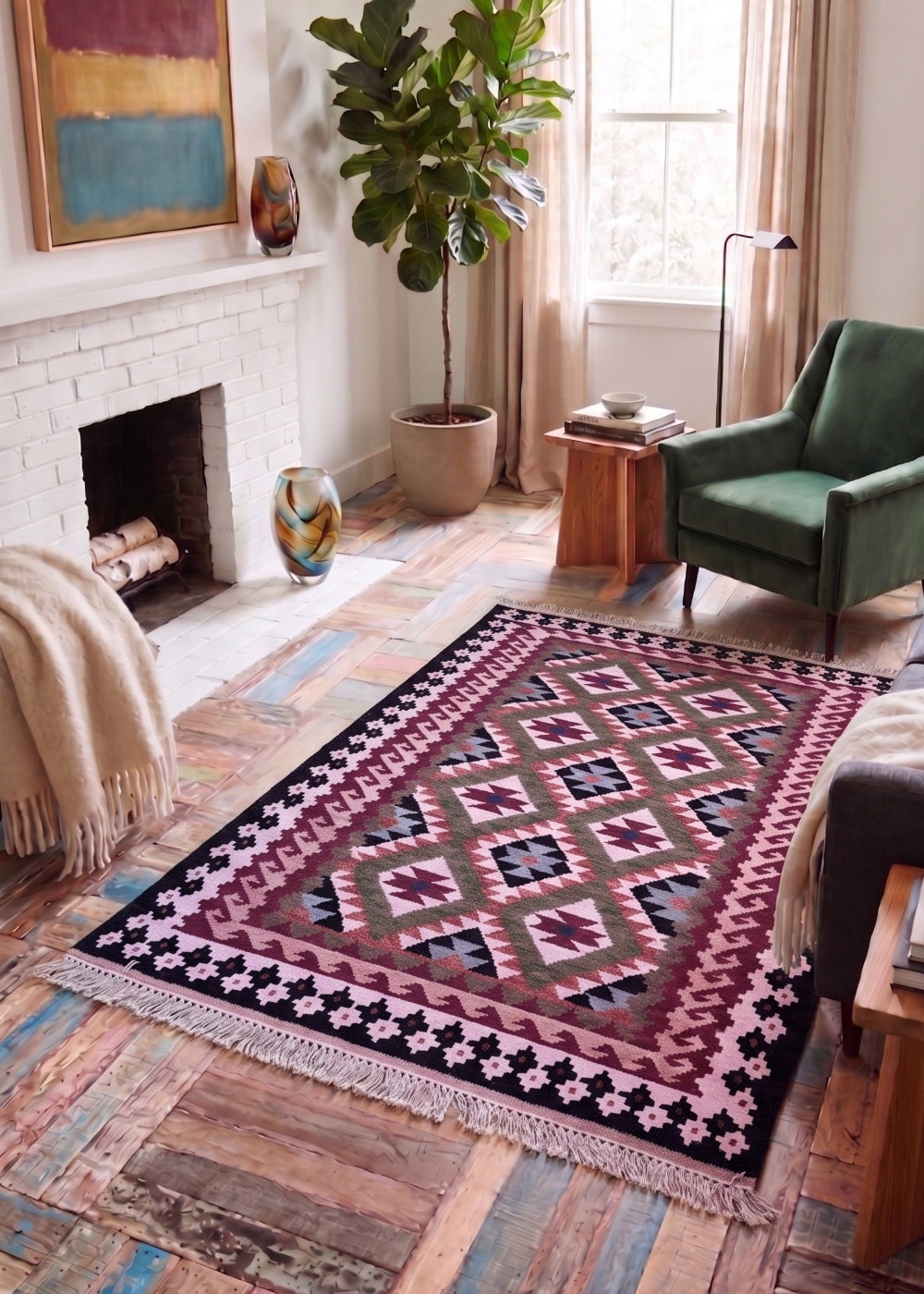 A rectangular area rug with a geometric Southwestern-style diamond pattern in pink, red, black, and green, styled on a rustic wooden floor in a cosy living room with a green armchair, fireplace, and potted plant.