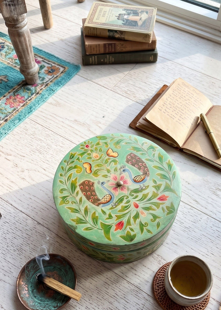 Decorative round box with nature-themed design on a wooden floor with books and a cup.